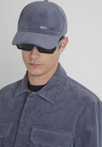 Giovane uomo che indossa un cappello di velluto a coste grigio e una giacca con tasche frontali, abbinati a occhiali da sole neri e una camicia nera sotto.