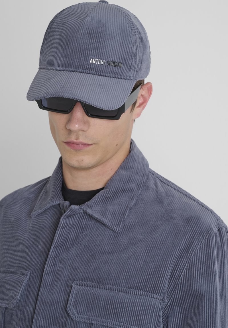 Giovane uomo che indossa un cappello di velluto a coste grigio e una giacca con tasche frontali, abbinati a occhiali da sole neri e una camicia nera sotto.