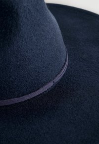 Marine blauwe vilten hoed met een brede rand, voorzien van een gladde band rond de kroon en een zachte textuur, die de gestructureerde vorm benadrukt.