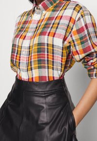 Woman wearing a multicolored plaid button-up shirt tucked into a high-waisted black leather skirt with hands in pockets.