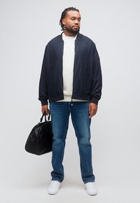 Άνδρας με πλεξούδες, ντυμένος με μαύρο μπουφάν, λευκό πουκάμισο, μπλε τζιν, λευκά αθλητικά παπούτσια, κρατώντας μαύρη δερμάτινη τσάντα, στέκεται μπροστά σε μονόχρωμο φόντο.