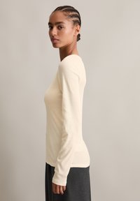 Langärmliges cremefarbenes Oberteil aus weichem, geripptem Material. Rundhalsausschnitt mit figurbetontem Schnitt, kombiniert mit einem dunkelgrauen Rock, der einen Texturkontrast zeigt.