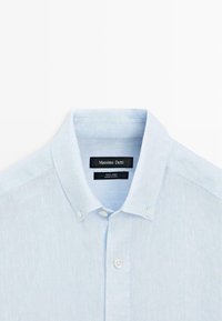 Light blue linen button-down shirt with a folded collar and white buttons, featuring a black label inside the neckline.