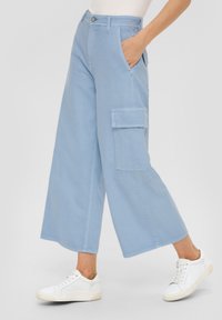 Hellblaue Cargo-Hosen aus Baumwolle. Sie haben weite Beine, Fronttaschen und eine Seitentasche mit Klappe. Kombiniert mit weißen Sneakern.