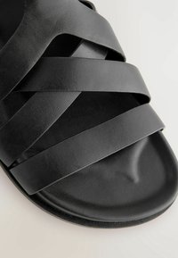 Gros plan d'une sandale noire avec trois larges brides croisées sur une semelle matelassée.