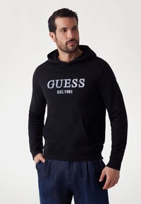 Sudadera negra con capucha de algodón, que presenta un logotipo bordado "GUESS EST.1981" en gris, bolsillos estilo canguro y puños de canalé.