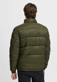 Olivenfarget dunjakke med høy krage, quilted design, elastiske mansjetter og en glatt tekstur. Bæres over svarte bukser.