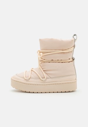 Puffer ankle boot in light beige, featuring elastic laces, a textured sole, and metallic accents along the back. Smooth, padded fabric construction.