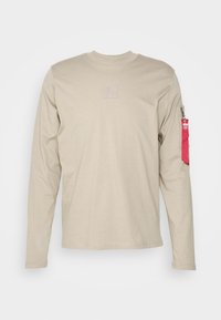Beige pitkähihaine paita, jossa on pyöreä pääntie, pienen kohokuvioidun logon rintaosassa ja punainen vetoketjullinen tasku vasemmassa hihassa.