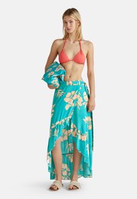 Korallenfarbener Bikini-Oberteil kombiniert mit einem türkisfarbenen Maxirock mit floralem Muster und einem fransigen Saum. Das Model trägt helle Slide-Sandalen.