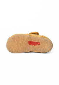 Scarpa di stoffa gialla con suola in gomma testurizzata. La suola presenta un motivo ondulato e un logo rosso, con punta rotonda e cinturino in velcro.