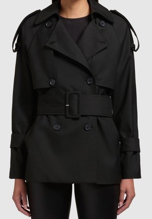 Mujer que lleva un abrigo negro doble botonadura con un cinturón ancho y hombreras abotonadas sobre pantalones negros.