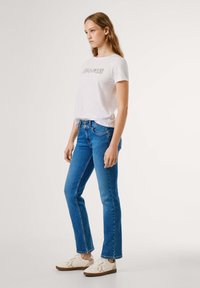 Ung kvinne står iført en hvit t-skjorte med grafikk, blå høytlivsjeans med dobbel knapp, og hvite sneakers med leoparddetalj.