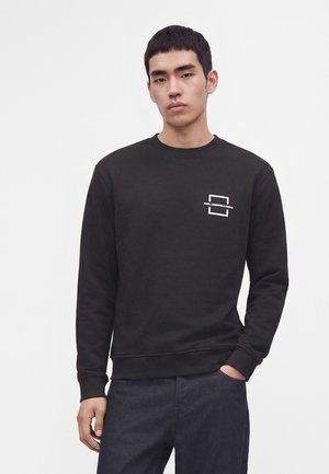 Joven con sudadera negra de manga larga con un pequeño logo blanco en el pecho y pantalones oscuros, de pie frente a un fondo liso.