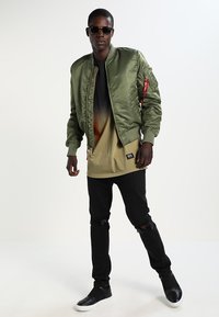 Green satin bomber jacket with ribbed collar, zip pockets, and red branding detail. Worn over an ombre gradient t-shirt and black distressed jeans.