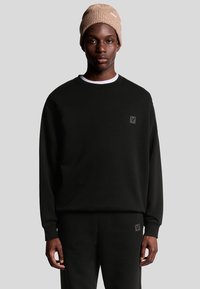 Felpa nera con maniche lunghe, colletto a giro, e un patch logo sul petto, abbinata a pantaloni neri coordinati e un berretto in maglia beige.