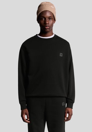 Schwarzes Sweatshirt mit langen Ärmeln, Rundhalsausschnitt und Logopatch auf der Brust, kombiniert mit passenden schwarzen Hosen und einer hellbraunen Strickmütze.