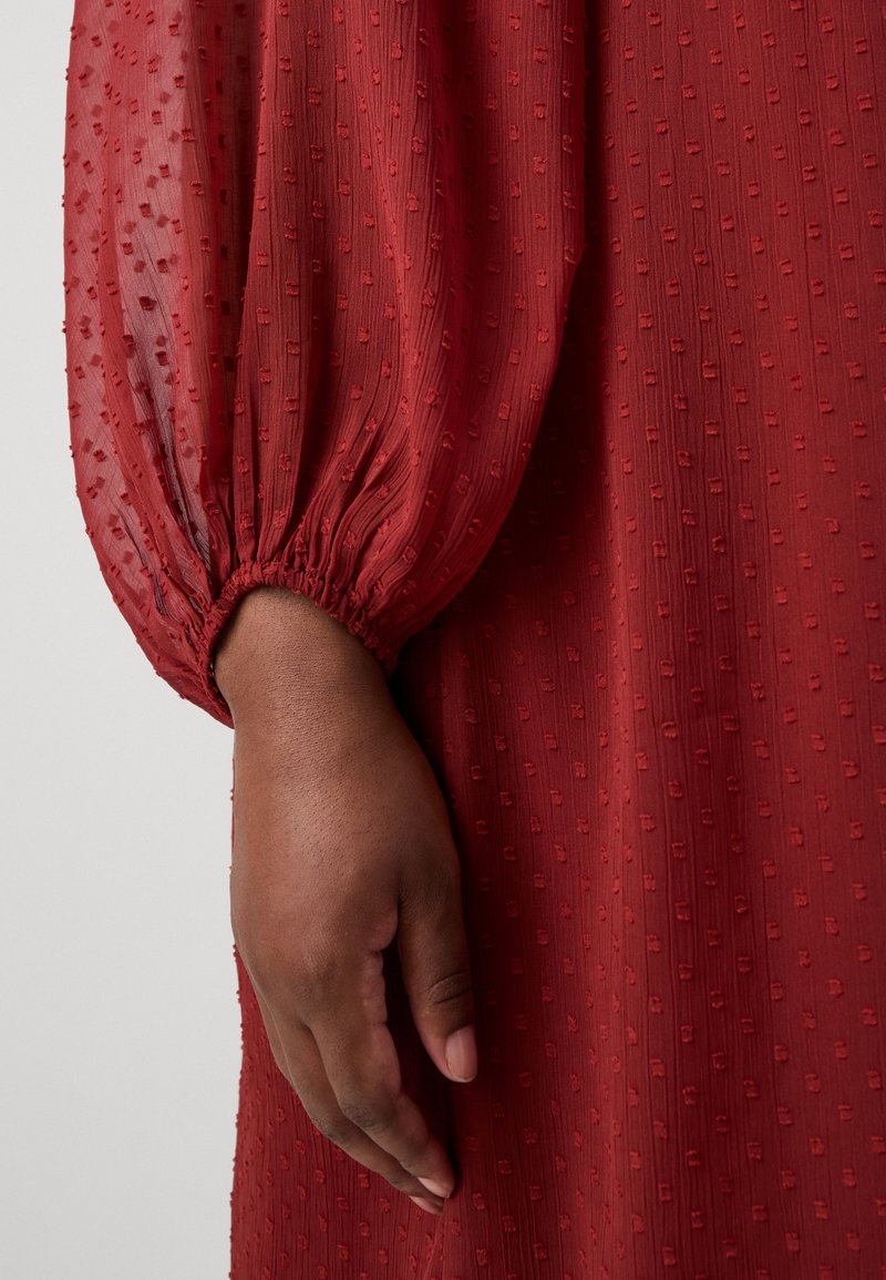 Rotes, strukturiertes Kleid mit Puffärmeln und gerafftem Bündchen. Der Stoff hat dezente, erhabene Muster, die Detail und Dimension verleihen.