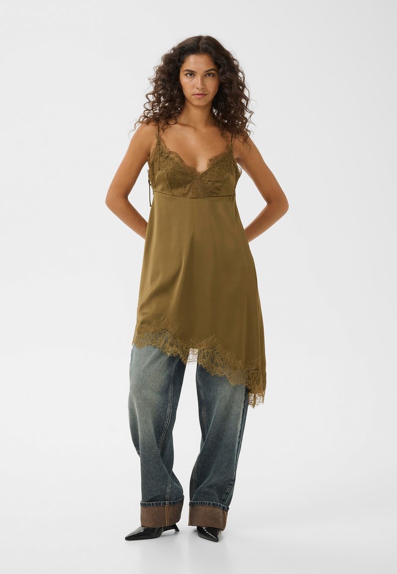 Femme aux cheveux bouclés portant une robe nuisette vert olive bordée de dentelle sur un jean large retroussé et des chaussures pointues noires, debout les mains dans le dos.
