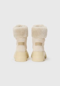 Búza színű bokacsizma, puha szőrmével a gallérján, texturált velúr felsőrésszel, dombornyomott logóval és vastag, tömzsi gumitalppal.