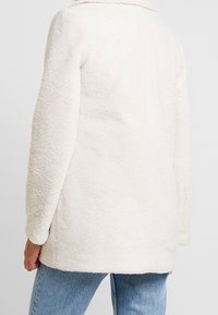 Jasno beżowy płaszcz z polaru o teksturowanej powierzchni, z kołnierzem, długimi rękawami i prostym krojem, zestawiony z jasnoniebieskimi dżinsami.