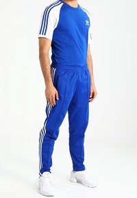 Mies, joka käyttää sinivalkoisia Adidas-urheiluvaatteita, joissa on kolme raitaa hihoissa ja housuissa, seisoo kädet rentoina sivuilla.