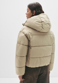 Giacca imbottita beige con cappuccio, caratterizzata da un design trapuntato e cuciture a coste. Il materiale appare liscio e leggermente lucido.
