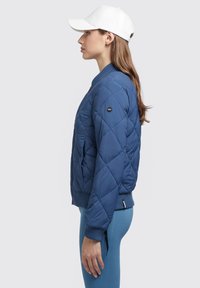 Quiltsētā tumši zilā bomber jaka ar ribotām aprocēm un apakšmalu, ar melnu logotipa uzšuvumu uz piedurknes, valkāta kopā ar zilām teksturētām legingām.