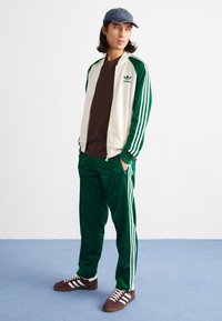 Tuta verde con strisce bianche, giacca crema con cerniera, maglia marrone e sneakers marroni con dettagli bianchi. Cappellino blu scuro.