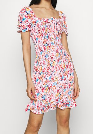 Kobieta w dopasowanej, krótkiej sukience w kwiaty z bufkami, kwadratowym dekoltem i marszczonym dołem w kolorach różowym, niebieskim, pomarańczowym i białym.