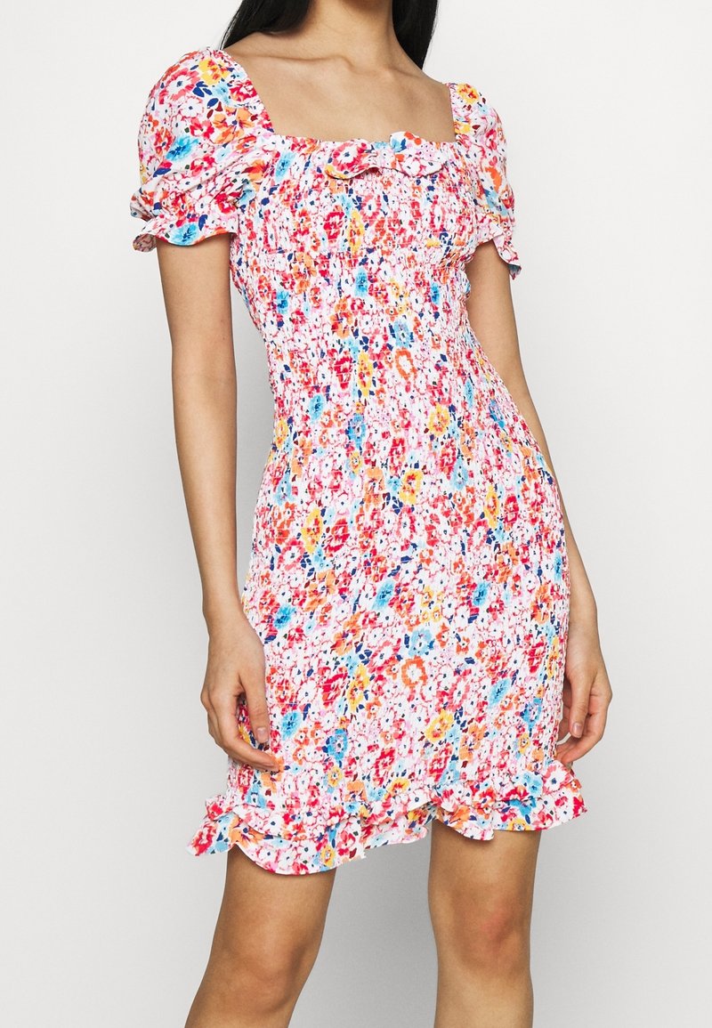 Femme portant une robe courte ajustée à fleurs avec des manches bouffantes, un décolleté carré et un ourlet à volants dans des couleurs rose, bleu, orange et blanc.