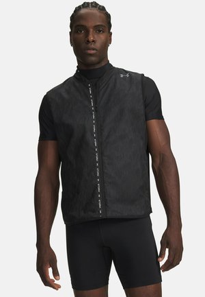 Homme aux cheveux tressés portant une veste noire sans manches zippée sur un haut de sport noir à manches courtes, et un short noir moulant, debout face à l'avant.