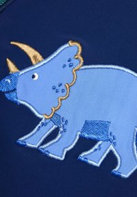 Kék hímzett triceratops sötétkék szövet háttéren, aranyszarv kiemelésekkel és textúrált részletezéssel a dinoszaurusz mintázatában.