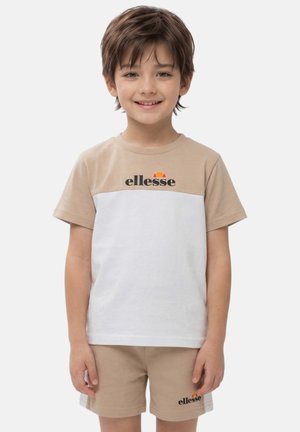 Bambino sorridente con capelli castani corti che indossa una t-shirt ellesse beige e bianca e pantaloncini beige abbinati, in piedi davanti a uno sfondo semplice.