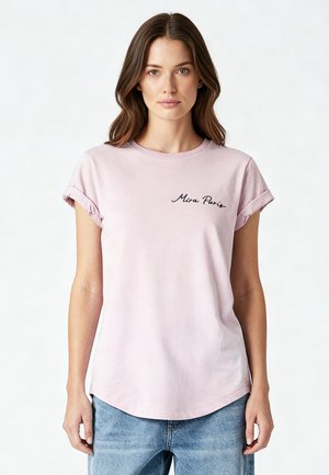 Donna che indossa una maglietta rosa chiaro con le maniche arrotolate e la scritta "Mira Paris" sul petto e jeans blu, in piedi davanti a uno sfondo bianco semplice.