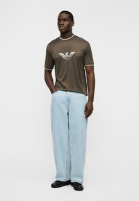 Άνδρας με καφέ κοντομάνικη μπλούζα με λευκό λογότυπο, ανοιχτό μπλε τζιν παντελόνι με φαρδιά μπατζάκια και μαύρα παπούτσια, στέκεται με το ένα χέρι στην τσέπη.