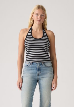 Femme blonde portant des boucles d'oreilles créoles, un haut à rayures noires et blanches à col halter, et un jean taille haute bleu clair, debout sur un fond uni.