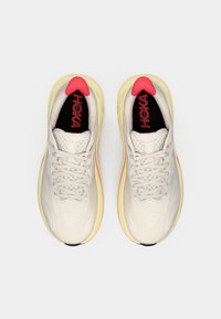 Scarpe da running color crema chiaro con una tomaia testurizzata, dettagli rossi a contrasto sulla linguetta e una suola gialla imbottita. Logo del marchio in nero e rosso visibile.