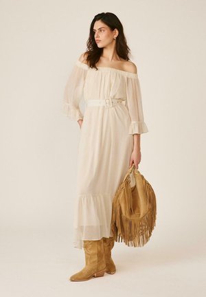 Mujer con vestido blanco sin hombros con cinturón, bolso marrón con flecos y botas vaqueras marrones, mirando hacia su izquierda contra un fondo liso.