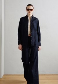 Donkere spijkerbroek met wijde pijpen, gecombineerd met een bijpassend donker overhemd bovenop een lichte top. Oversized zonnebril maakt de look af.