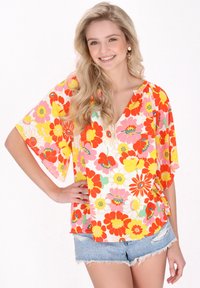 Blusa floreale con maniche corte e larghe nei toni del rosa, giallo e rosso; presenta un col V e una vestibilità comoda, abbinata a pantaloni corti in denim.