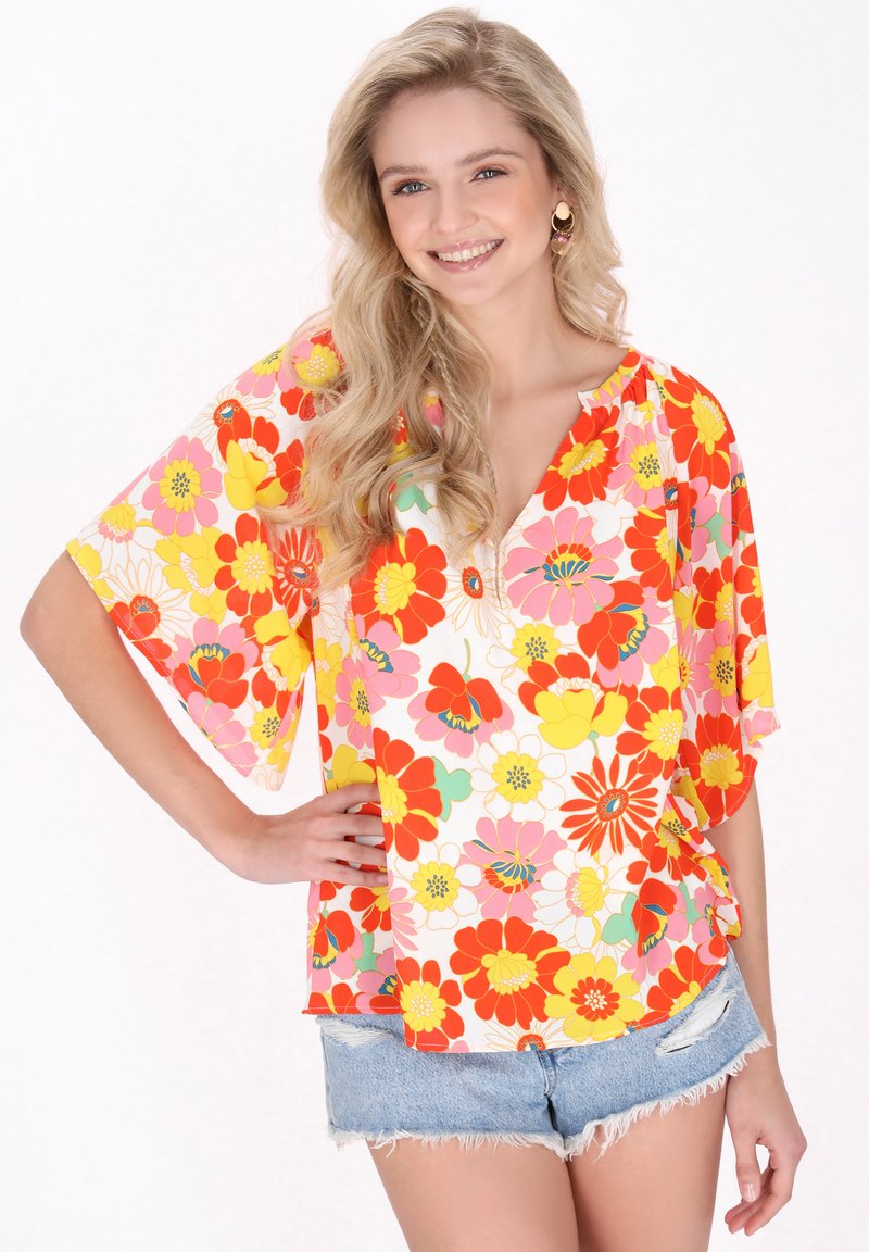 Blusa floreale con maniche corte e larghe nei toni del rosa, giallo e rosso; presenta un col V e una vestibilità comoda, abbinata a pantaloni corti in denim.