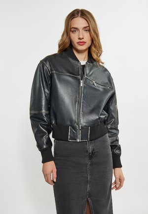 Giubbotto bomber in pelle nera con polsini e orlo a coste, dotato di chiusura zip sul davanti e tasche laterali, abbinato a una gonna di jeans scura.