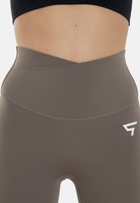 Hoge taille legging in een gedempte bruin tint, met een kruisdessign aan de tailleband en een wit logo accent aan de zijkant.