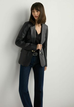 Kobieta ubrana w czarną skórzaną marynarkę, czarny top i ciemnoniebieskie dżinsy, stojąca na tle jasnej, gładkiej ściany.