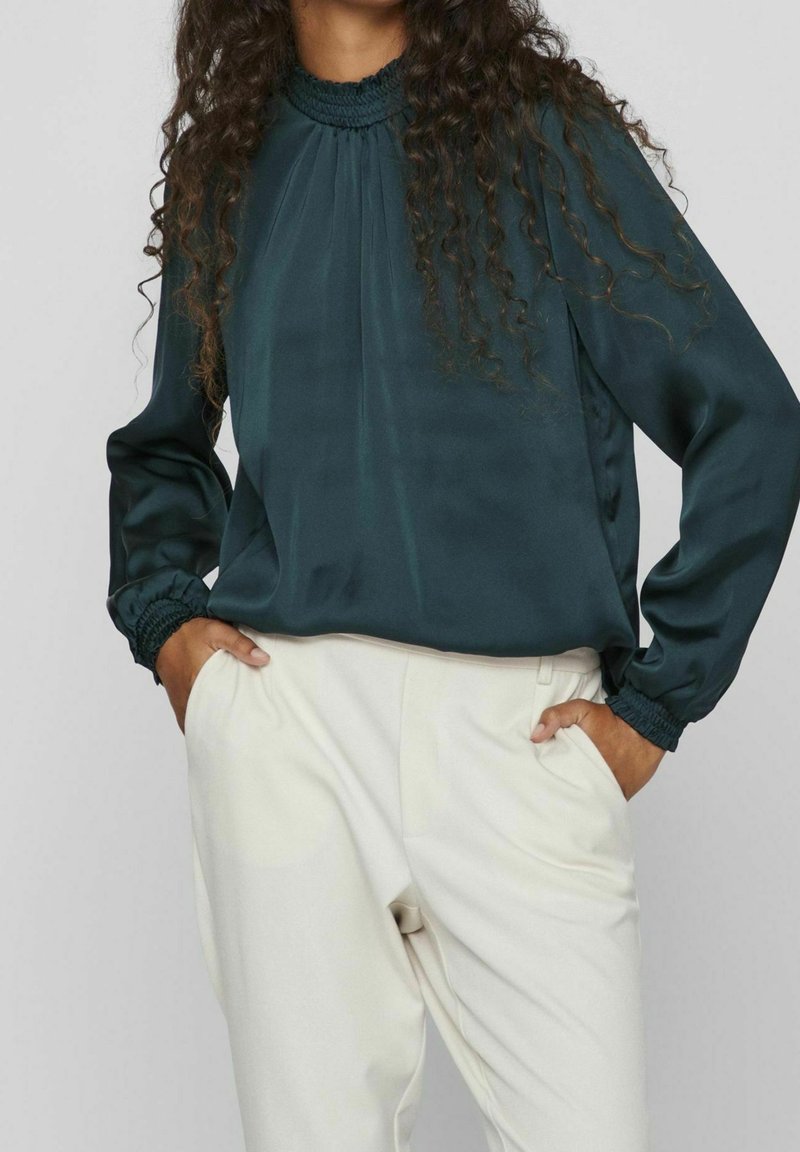 Bluzka z satyny w kolorze morskim, z długimi rękawami, marszczonym dekoltem i elastycznymi mankietami, w połączeniu z kremowymi spodniami, które mają boczne kieszenie i gładką fakturę.