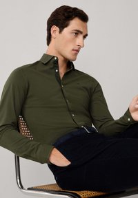 Camisa de manga larga color verde oliva oscuro con un collar negro en contraste, diseño ajustado, tapeta de botones y un sutil logotipo cerca del pecho.
