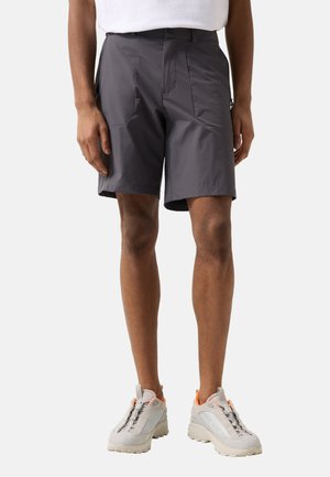 Mann, der dunkelgraue knielange Shorts und weiße Turnschuhe trägt, steht vor einem schlichten Hintergrund.