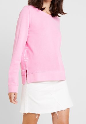 Kvinna som bär en långärmad rosa tröja med sidospjälor och en vit minikjol med fransad fåll, visas från halsen till mitten av låret.