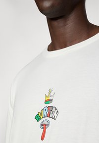 Witte katoenen t-shirt met een kleurrijk grafisch ontwerp met een kroon, het woord "BURTON," en een cartooneske tong, tegen een effen achtergrond.